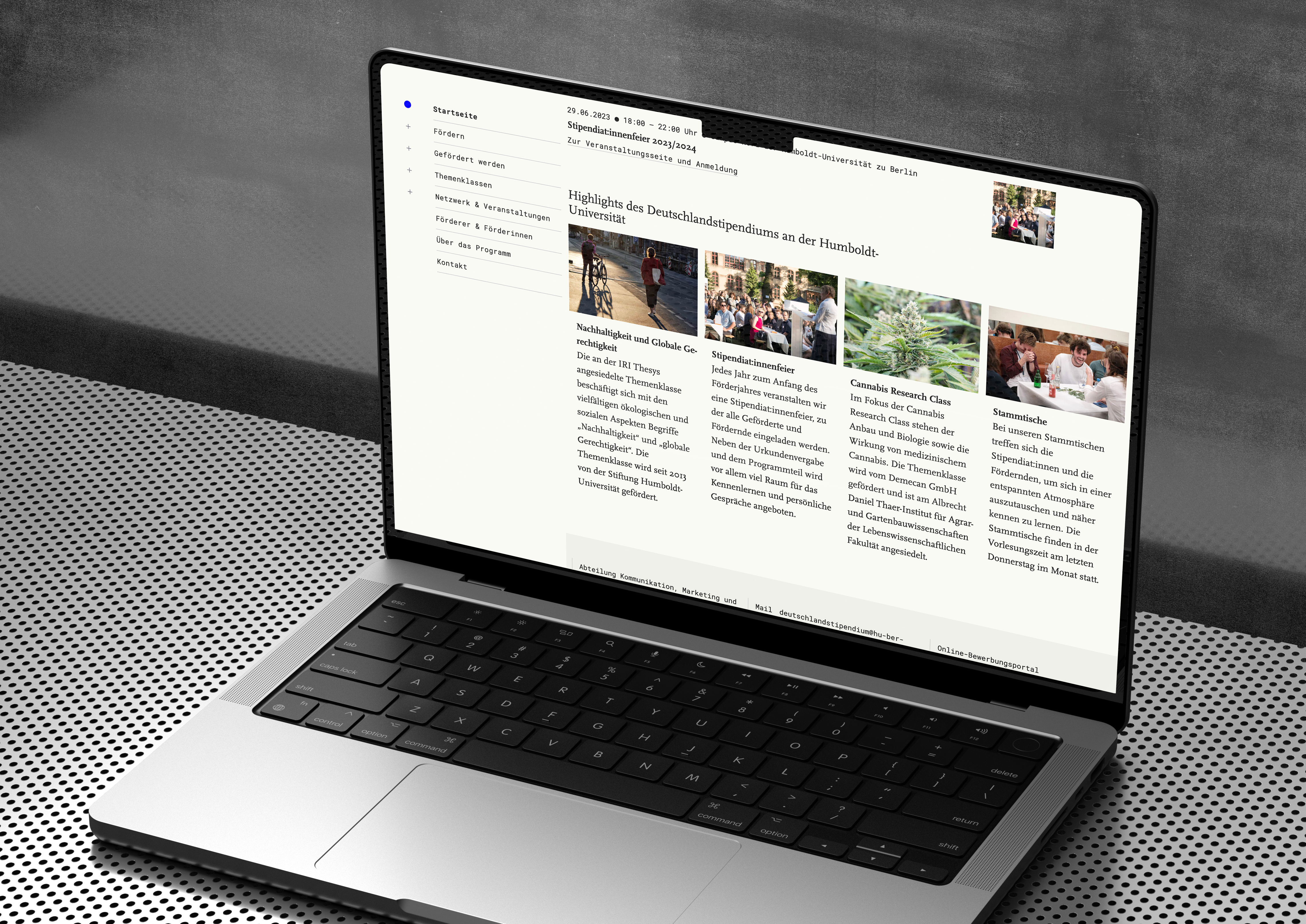 A macbook pro on a perforated metal plate with a concrete wall in the back. The screen shows a part of a website with a lot of light background and different navigation tiles. The look is clean and elegant.