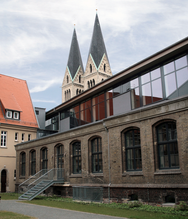 A photo of the campus Halberstadt. There is a historical building in the foreground with a glass front additional story on top. In the background you can see two church towers that give the impression of a historical city center.
