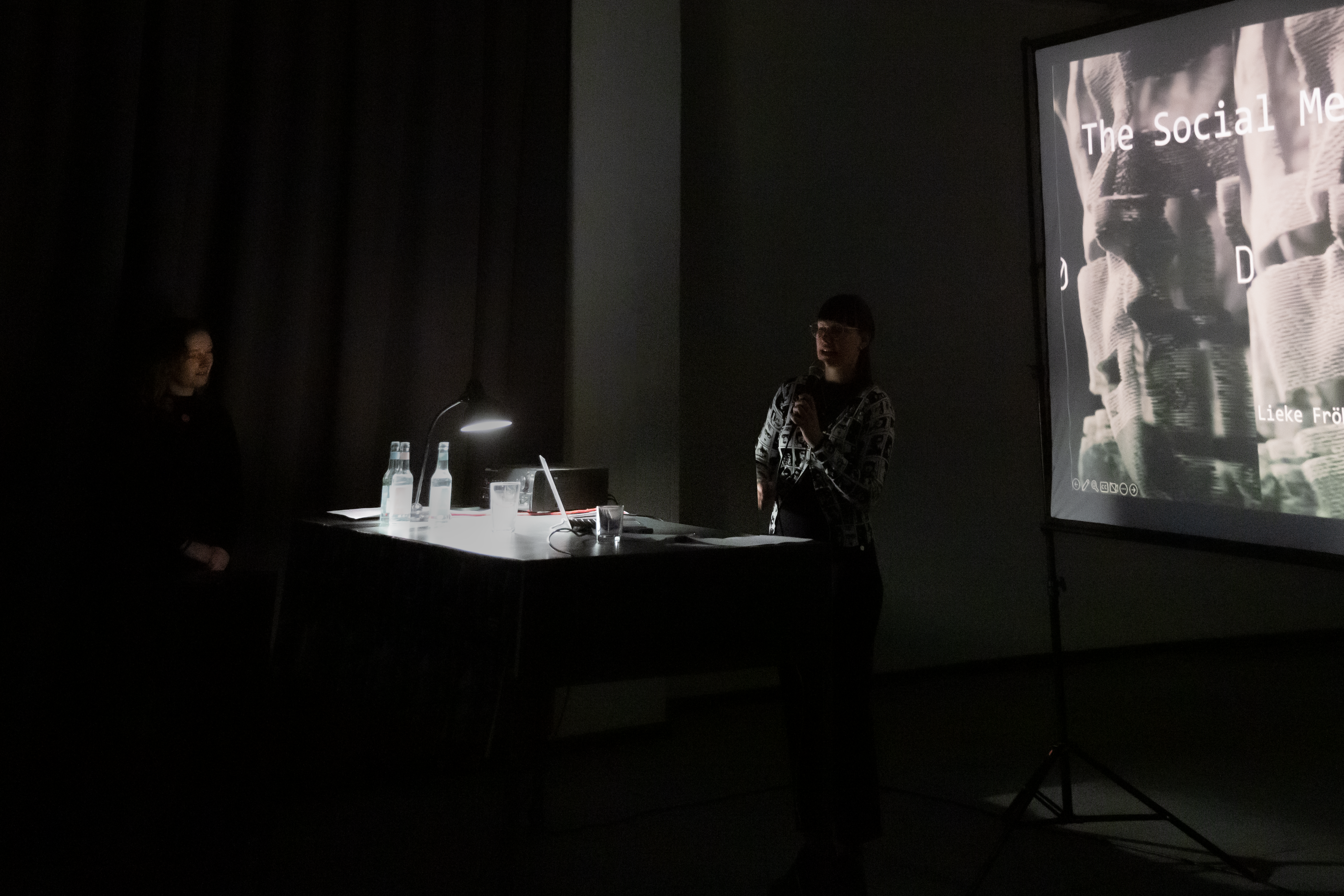The speaker of a talk in front of the screen. The room is dark, only the table is lit by a spotlight lamp. A person standing on the left watching the speaker.