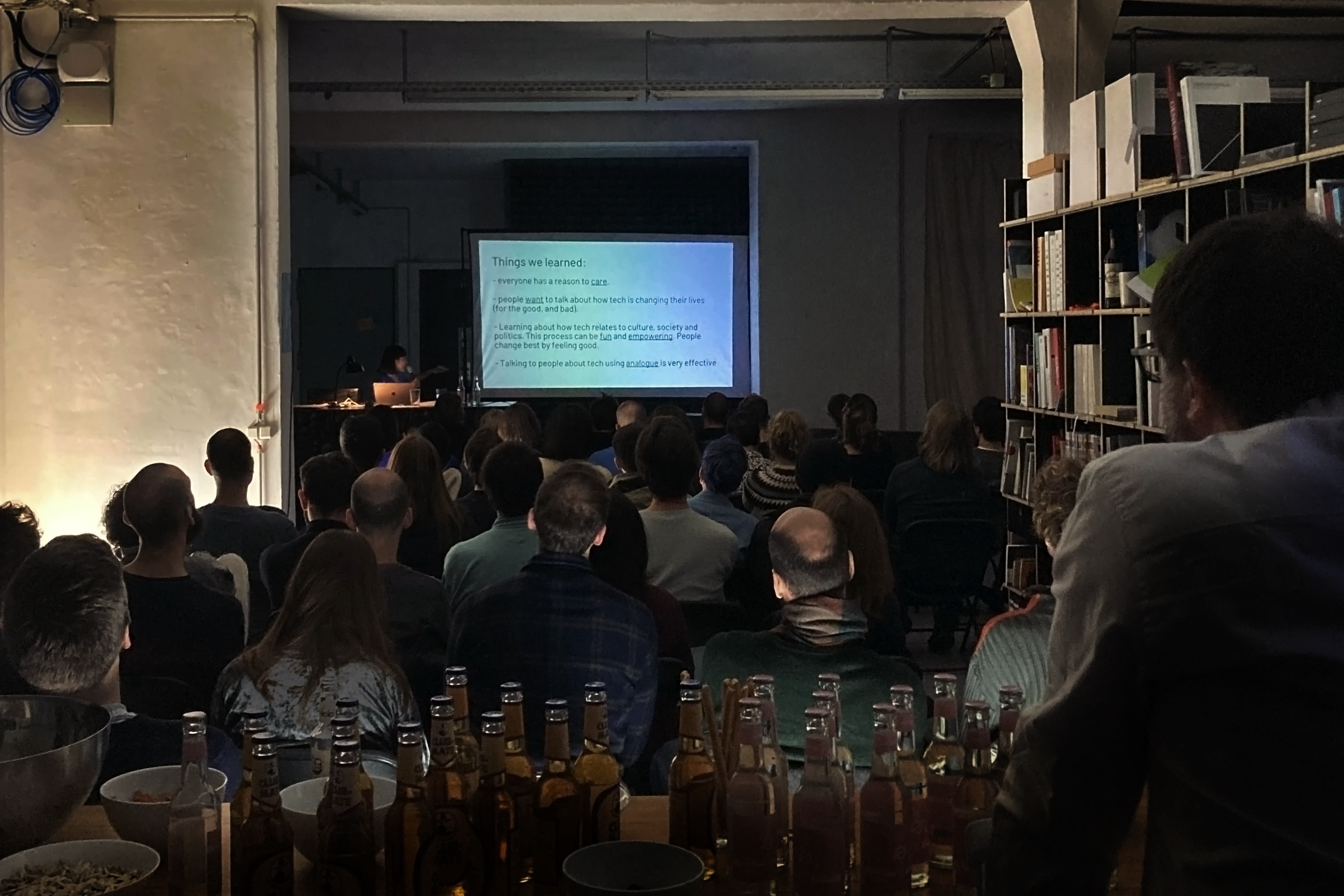 A talk in a design studio. The view on the speaker with screen in dim light in the back. The sitting audience of around 60 people from behind. The foreground is the back of a person.