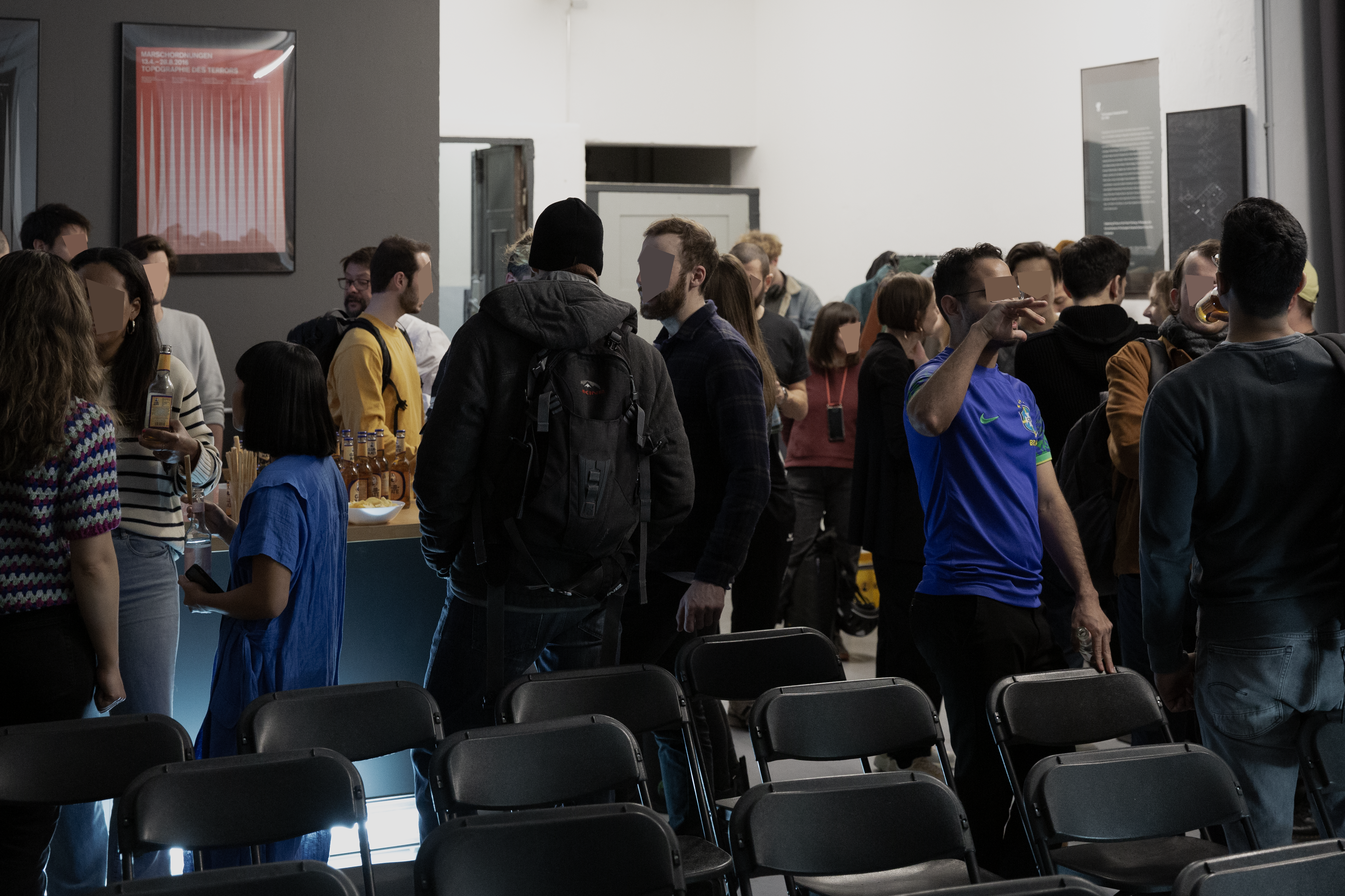 A crowd in a design studio. Empty chairs in the front, the kitchen and entrance space is tightly packed. People are talking, faces are anonymised.