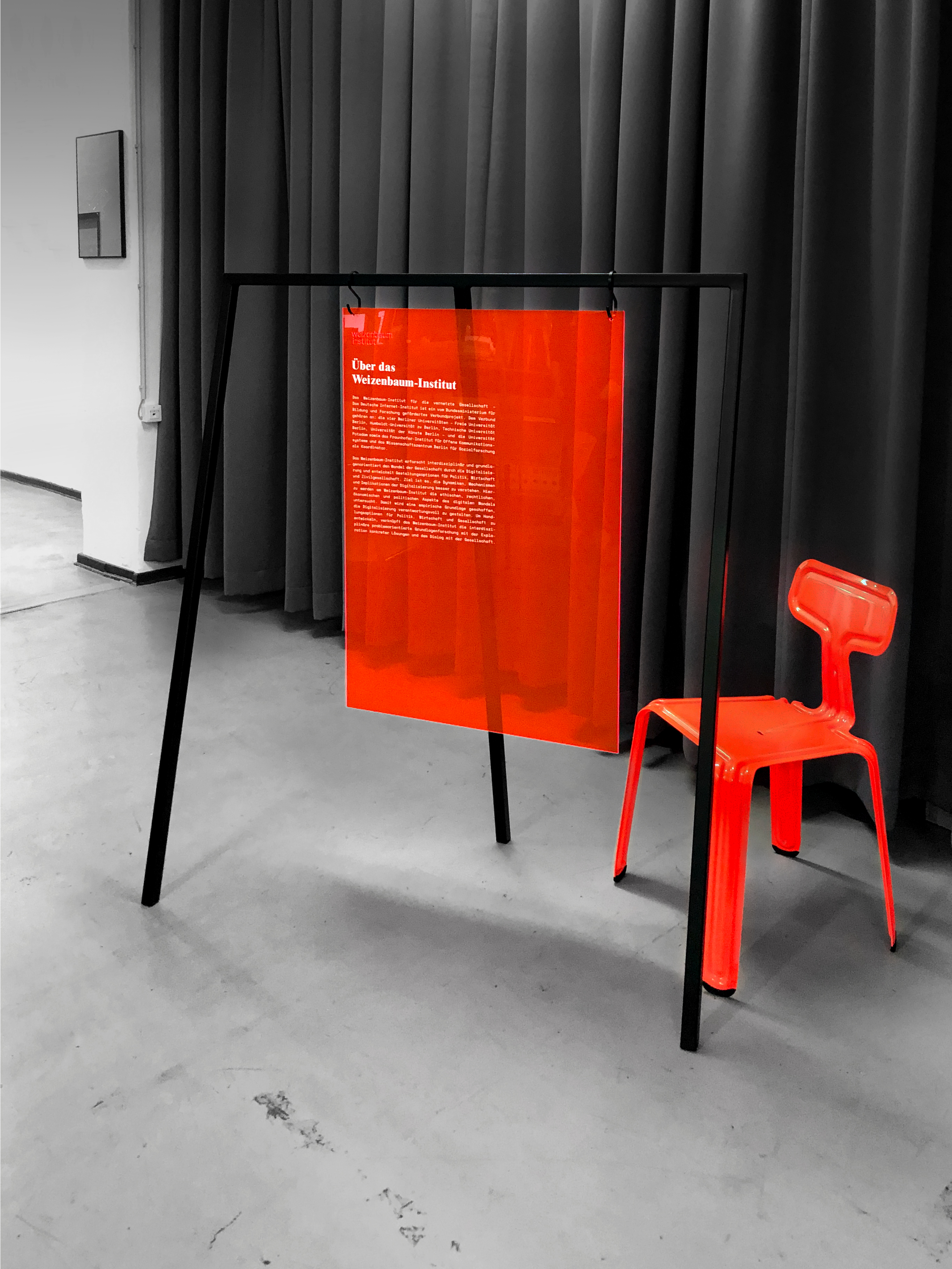 A photo showcasing a semi-transparent red acrylic poster hung on a metal frame and a red metallic chair to the right side, in the background a black curtain can be seen.